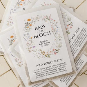 May include: A white card with a floral design and the text "Baby in Bloom" and "Angelica's Baby Shower 07.21.25". The card is attached to a small white packet of wildflower seeds. The packet has instructions for planting the seeds.