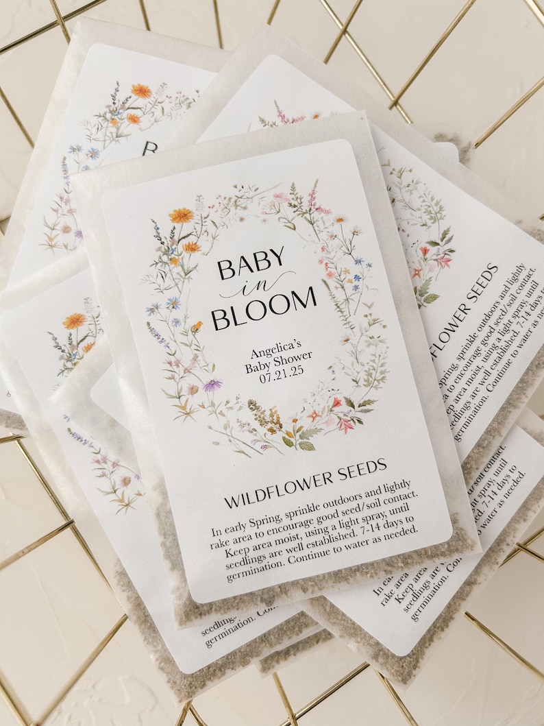 May include: A white card with a floral design and the text "Baby in Bloom" and "Angelica's Baby Shower 07.21.25". The card is attached to a small white packet of wildflower seeds. The packet has instructions for planting the seeds.