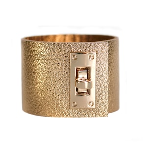 Può includere: Un ampio bracciale a polsino dorato con una superficie strutturata e una chiusura rettangolare. La chiusura è dorata e presenta una chiusura a cerniera. Il bracciale è realizzato in materiale metallico.