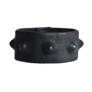 Puede incluir: Un brazalete de cuero negro con una superficie texturizada. El brazalete presenta tres detalles redondeados en relieve y está sobre un fondo blanco. El brazalete mide aproximadamente 5 cm de ancho.