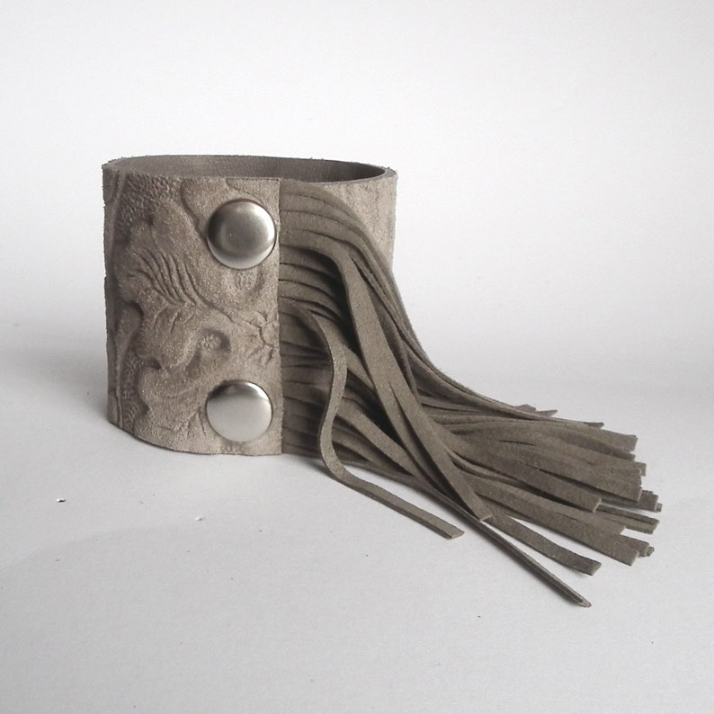 May include: A gray leather cuff bracelet with a fringe detail and two silver snaps.