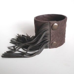 May include: A brown leather cuff bracelet with a fringe detail and two silver-toned snap closures.