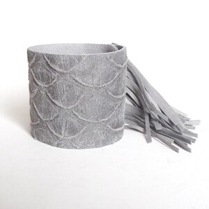 May include: A gray leather cuff bracelet with a fringe detail. The bracelet has a textured, embossed pattern.