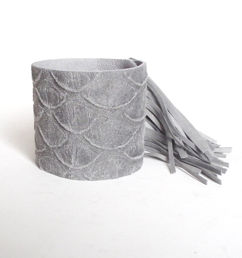 May include: A gray leather cuff bracelet with a fringe detail. The bracelet has a textured, embossed pattern.