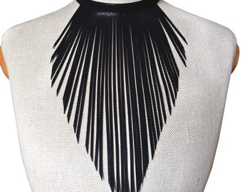 Puede incluir: Un collar gargantilla negro con un diseño de babero con flecos. El collar presenta una fina banda alrededor del cuello y una gran franja puntiaguda que se extiende por el pecho. La franja está hecha de tiras largas y finas.