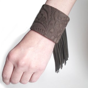 May include: A brown leather cuff bracelet with a floral embossed design and a fringe detail.