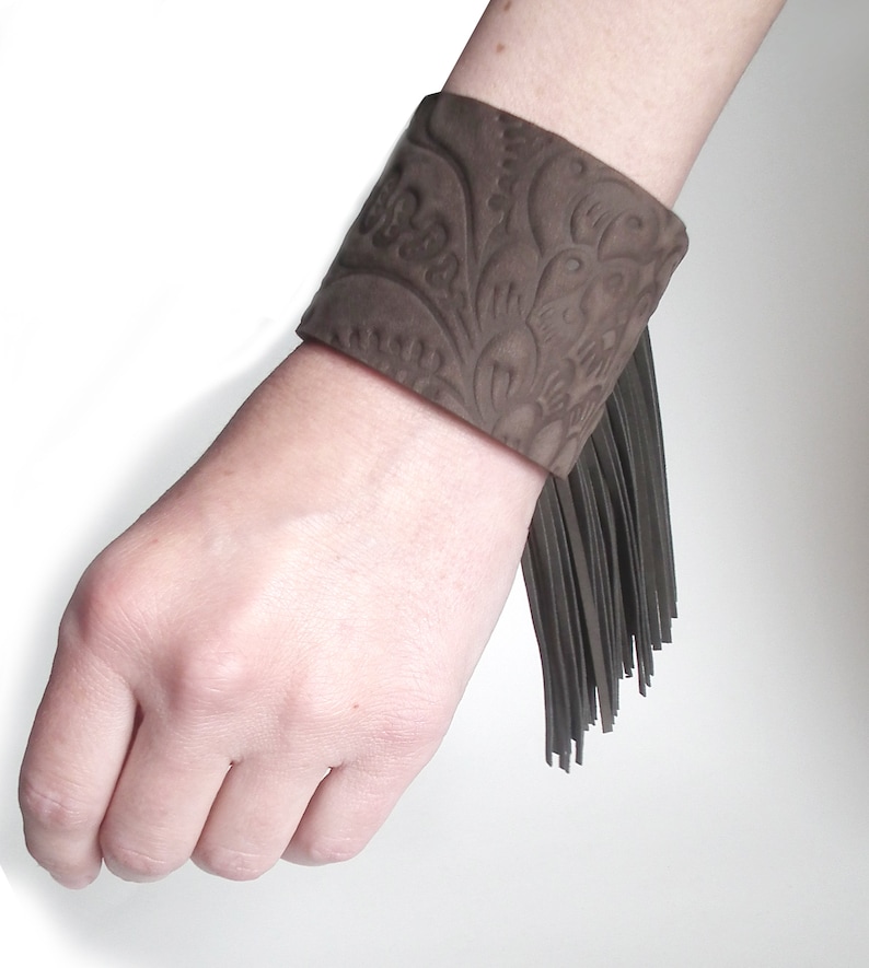 May include: A brown leather cuff bracelet with a floral embossed design and a fringe detail.