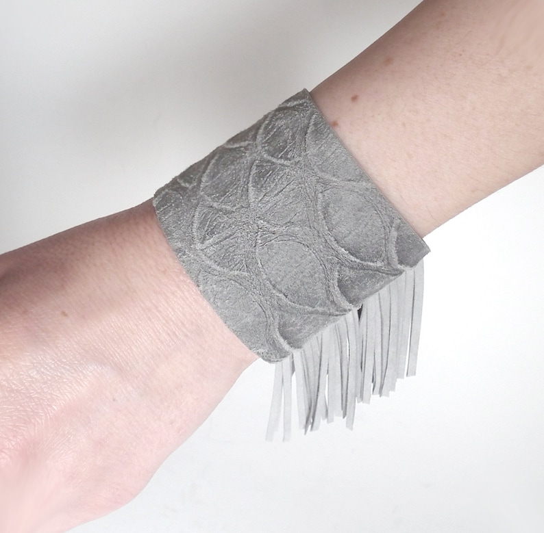May include: Gray suede bracelet with a fringe detail and a textured pattern.