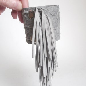 May include: Gray leather bracelet with a long fringe. The bracelet has two silver snaps.