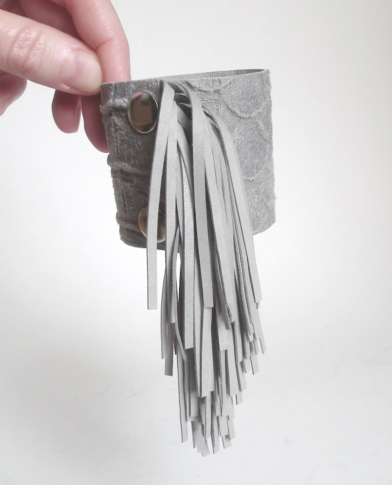 May include: Gray leather bracelet with a long fringe. The bracelet has two silver snaps.