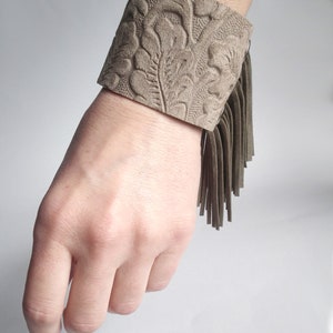 May include: A brown leather cuff bracelet with a floral embossed design and a fringe detail.