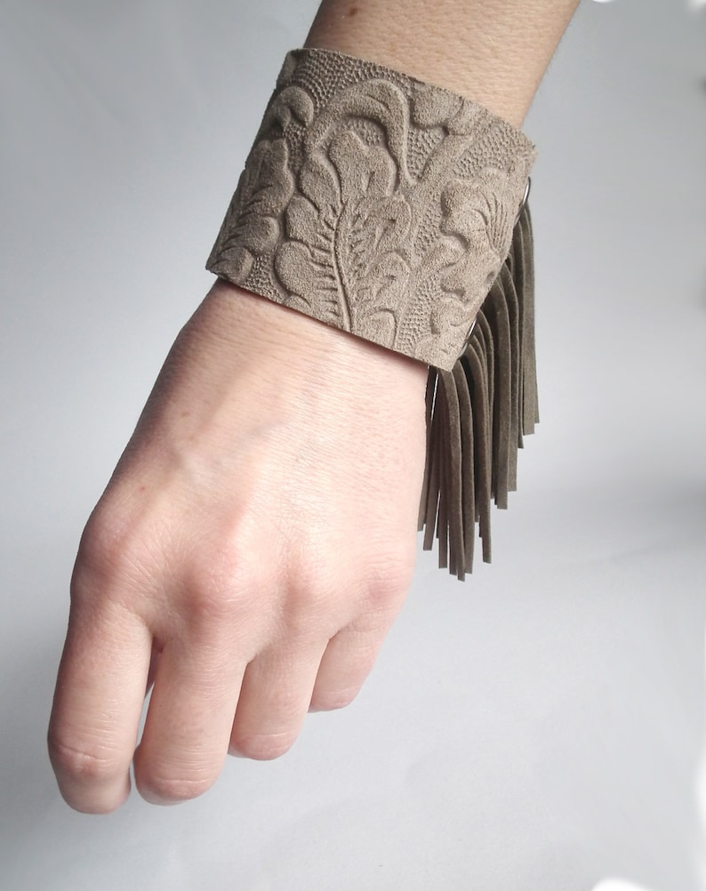 May include: A brown leather cuff bracelet with a floral embossed design and a fringe detail.