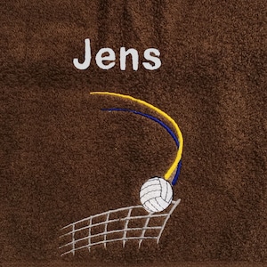 Könnte beinhalten: Braunes Handtuch mit dem Namen "Jens" in weiß gestickt. Ein weißer Volleyball wird über ein graues Netz mit blauen und gelben Linien serviert.