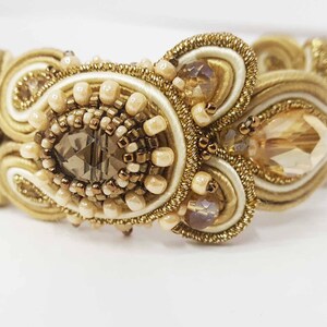 May include: A gold-toned bracelet with intricate beadwork and a central, faceted, smoky-quartz-colored stone. The bracelet features cream-colored and gold-colored woven elements, accented with small, round beads and larger, teardrop-shaped crystals. The design is ornate and elegant.