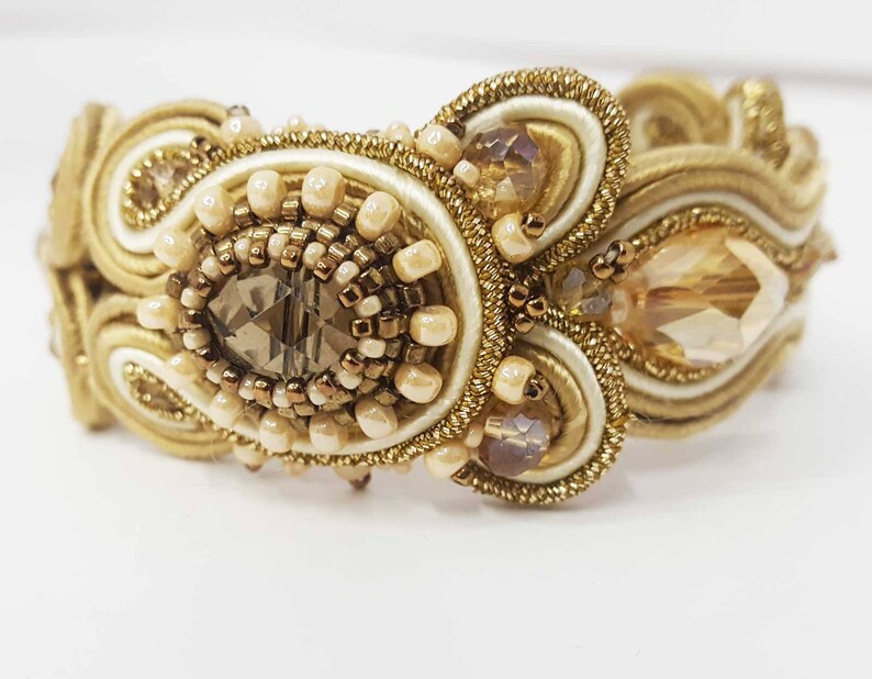 May include: A gold-toned bracelet with intricate beadwork and a central, faceted, smoky-quartz-colored stone. The bracelet features cream-colored and gold-colored woven elements, accented with small, round beads and larger, teardrop-shaped crystals. The design is ornate and elegant.