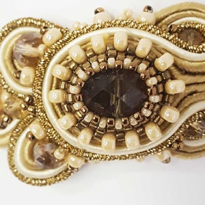May include: Close-up of an ornate, handcrafted jewelry piece. The design features a central dark brown, faceted gem surrounded by cream-colored beads and gold-toned accents. The piece is embellished with intricate gold threadwork and additional crystal-like embellishments.
