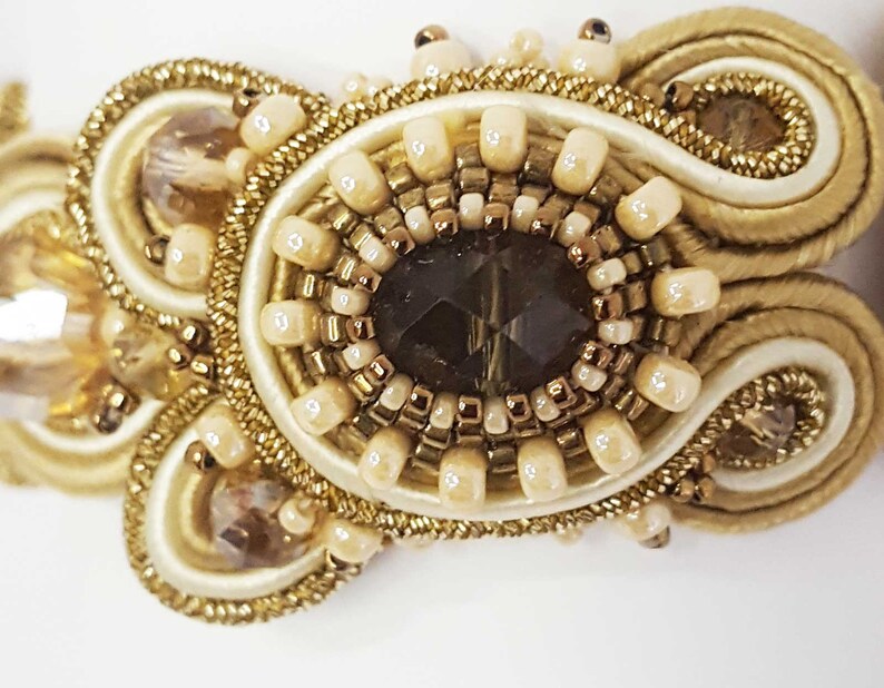May include: Close-up of an ornate, handcrafted jewelry piece. The design features a central dark brown, faceted gem surrounded by cream-colored beads and gold-toned accents. The piece is embellished with intricate gold threadwork and additional crystal-like embellishments.