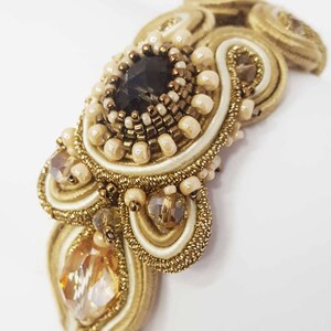 May include: Close-up of a handcrafted, ornate bracelet featuring a central dark gemstone surrounded by small beads and intricate gold-colored embroidery. The design incorporates cream-colored satin and sparkling crystals, creating a luxurious aesthetic.