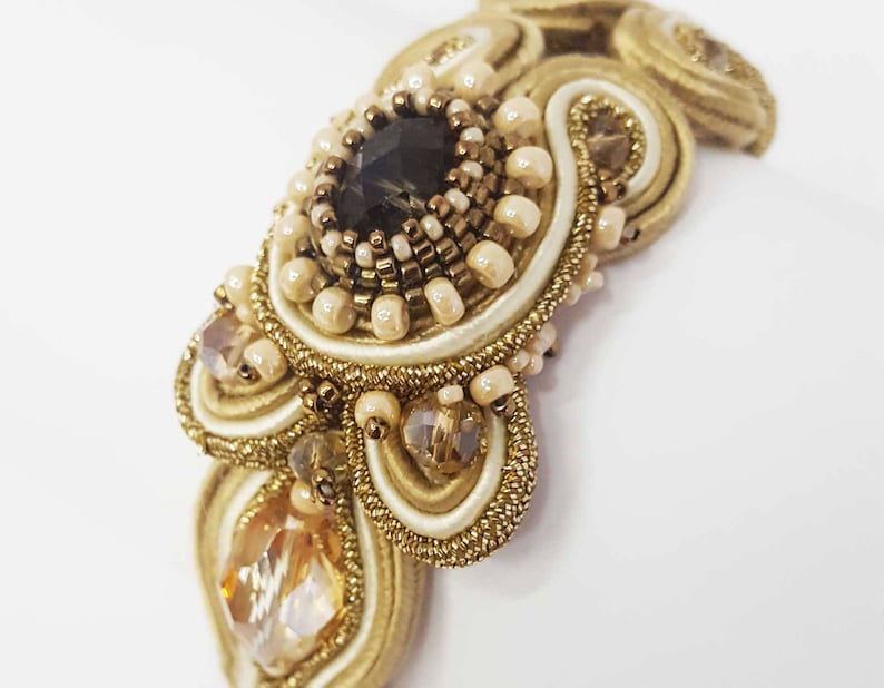May include: Close-up of a handcrafted, ornate bracelet featuring a central dark gemstone surrounded by small beads and intricate gold-colored embroidery. The design incorporates cream-colored satin and sparkling crystals, creating a luxurious aesthetic.