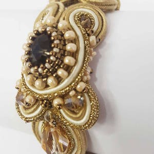 May include: A gold-toned bracelet with intricate beadwork and a large, dark central stone. The design incorporates cream-colored accents, small pearls, and sparkling crystals, creating a luxurious and ornate appearance. The bracelet is likely a statement piece.