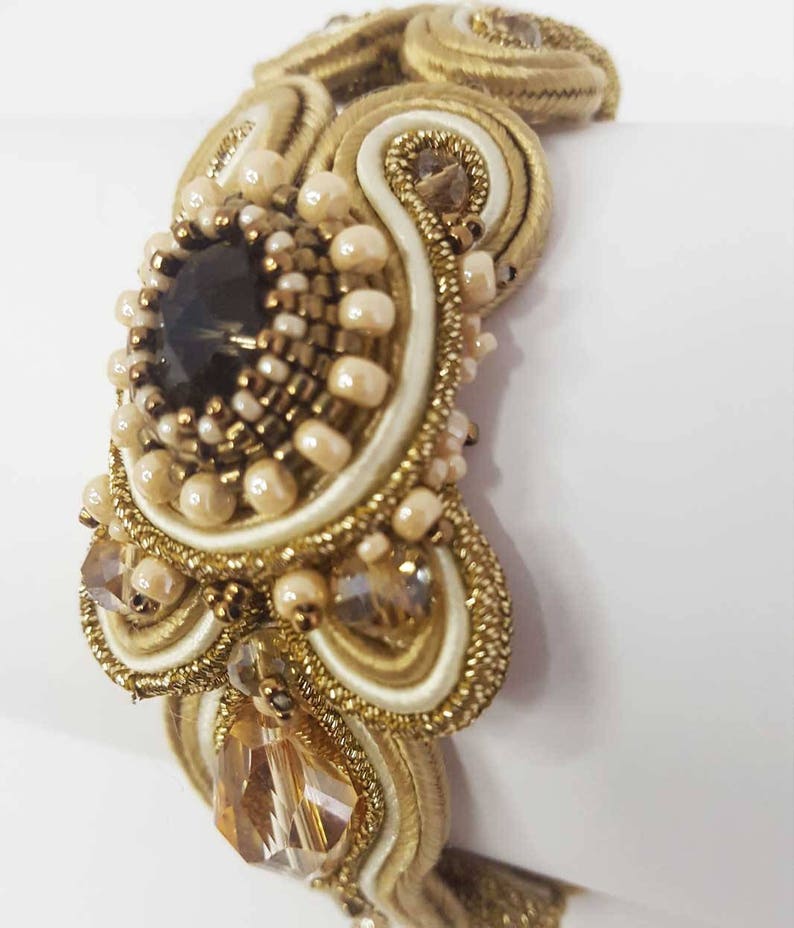 May include: A gold-toned bracelet with intricate beadwork and a large, dark central stone. The design incorporates cream-colored accents, small pearls, and sparkling crystals, creating a luxurious and ornate appearance. The bracelet is likely a statement piece.