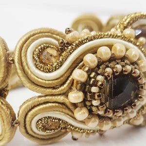 May include: Close-up of a gold-toned bracelet with intricate beadwork and a central dark-colored stone. The bracelet features cream-colored and gold-colored woven elements, with small, round beads and sparkling accents.