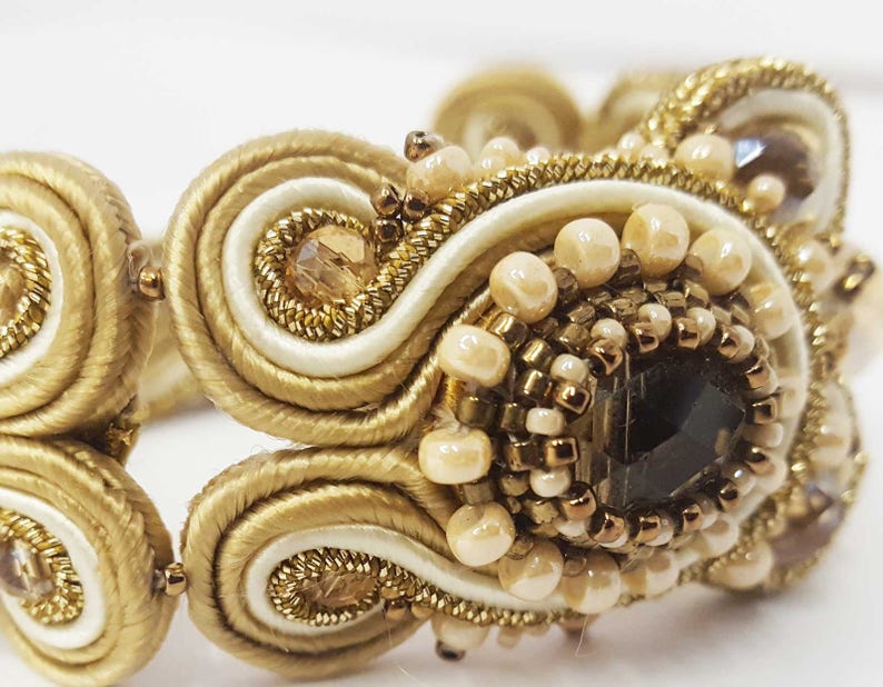 May include: Close-up of a gold-toned bracelet with intricate beadwork and a central dark-colored stone. The bracelet features cream-colored and gold-colored woven elements, with small, round beads and sparkling accents.