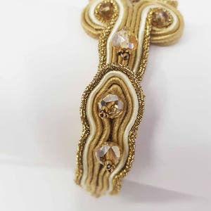 May include: Close-up of a gold and cream-colored bracelet with three clear, faceted gemstones. The bracelet features intricate woven detailing with gold thread and cream-colored satin ribbon. The gemstones are set in gold-colored metal.