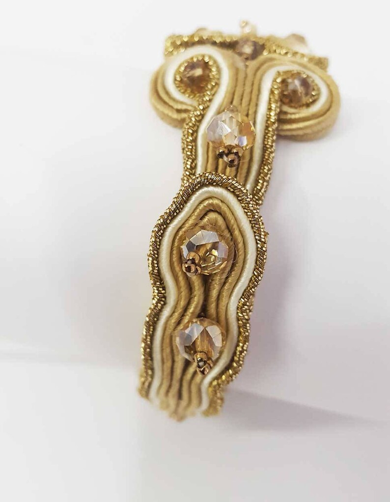 May include: Close-up of a gold and cream-colored bracelet with three clear, faceted gemstones. The bracelet features intricate woven detailing with gold thread and cream-colored satin ribbon. The gemstones are set in gold-colored metal.