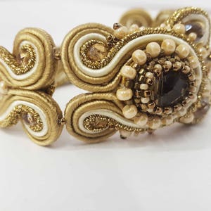 May include: A gold-toned bracelet with intricate detailing. The bracelet features a central dark gemstone surrounded by small, light-colored beads and gold thread. The design includes swirling patterns and a mix of textures.