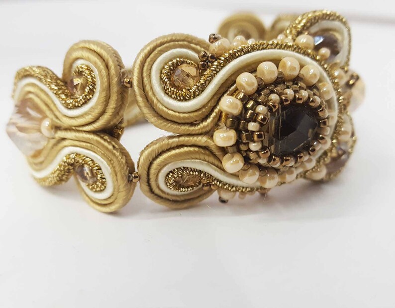 May include: A gold-toned bracelet with intricate detailing. The bracelet features a central dark gemstone surrounded by small, light-colored beads and gold thread. The design includes swirling patterns and a mix of textures.