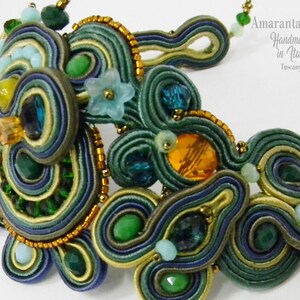 Può includere: Primo piano di una collana fatta a mano con intricato ricamo soutache. La collana presenta un design a spirale nei toni del verde, blu e oro, accentuato da perline e cristalli. Il testo "AmarantaBeads Handmade in Italy Tuscany" è visibile.