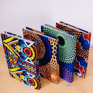 Könnte beinhalten: Vier Notizbücher mit Hardcover-Einbänden, die mit farbenfrohen afrikanischen Wachsdruckstoffen verziert sind. Die Einbände zeigen geometrische Muster in Gelb, Orange, Blau und Grün. Sie sind auf einer hellen Holzoberfläche angeordnet.