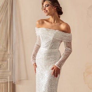May include: A white off-the-shoulder sequined wedding dress with long sleeves and a fitted silhouette. The dress has a train and a belt at the waist. The text "Lace Marry designs by Mary Zin & Gina Dung Hua" is printed on the bottom right corner of the image.