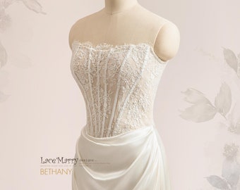 May include: A white strapless wedding dress with a lace bodice and a flowing satin skirt. The bodice features a corset-style design with boning and a sweetheart neckline. The skirt is gathered at the waist and falls to the floor in a soft, draping style. The dress is called "Bethany" by LaceMarry designs.