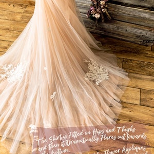 May include: A close-up of a blush-colored tulle wedding dress with delicate white lace appliques. The skirt is fitted on the hips and thighs and then flares out dramatically towards the bottom. The text "The Shirt is Fitted on Hips and Thighs and then it Dramatically Flares out towards the Bottom. It's finished by Flower Appliques and Lace Pieces scattered around in a Relaxed and Dreamy way!!!" is written on the dress.
