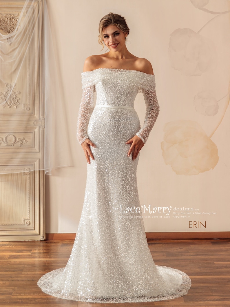 May include: A white off-the-shoulder wedding dress with long sleeves and a fitted silhouette. The dress is covered in sequins and has a satin sash at the waist. The dress is called "ERIN" and is designed by "Lace Marry".