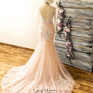 May include: A long-sleeved, blush-colored wedding dress with a lace bodice and a flowing tulle skirt. The dress has a deep V-neckline and a fitted silhouette. The dress is displayed on a mannequin against a rustic wooden background.