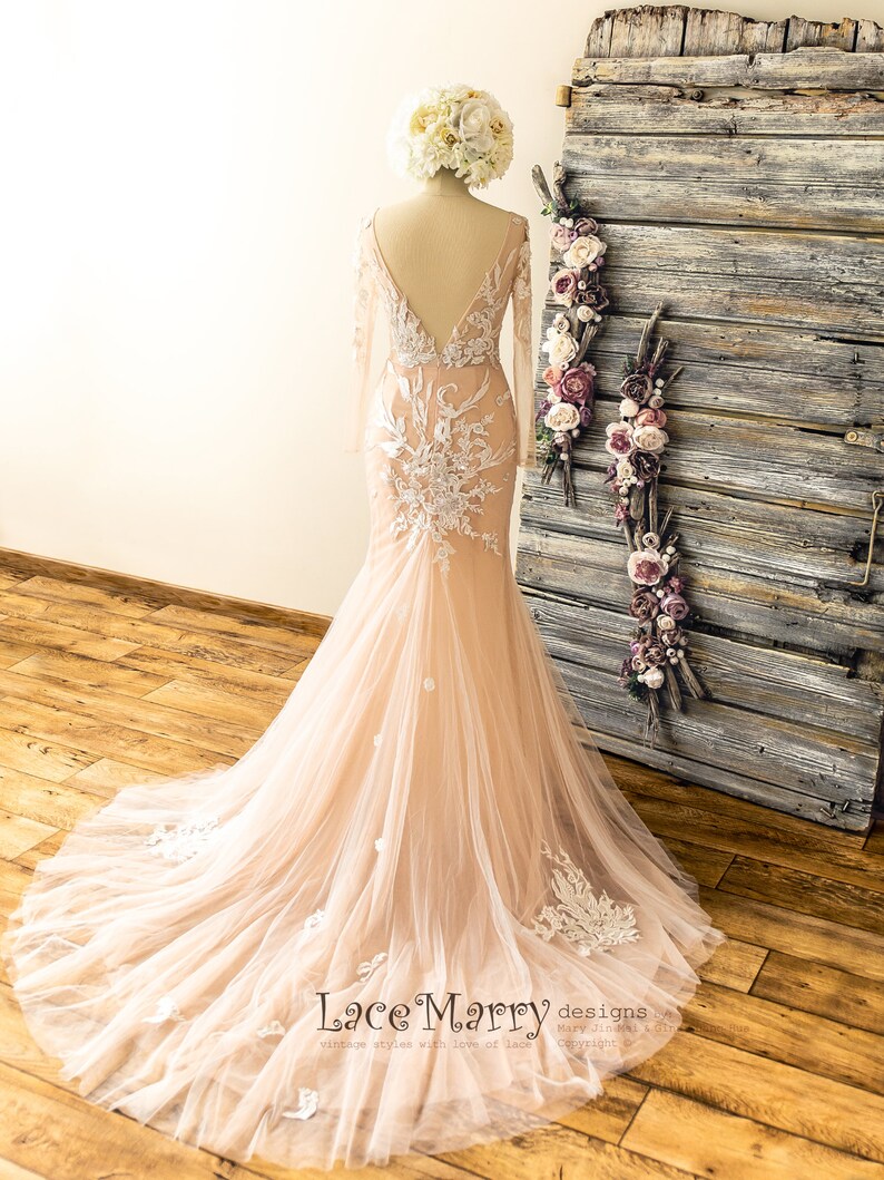 May include: A long-sleeved, blush-colored wedding dress with a lace bodice and a flowing tulle skirt. The dress has a deep V-neckline and a fitted silhouette. The dress is displayed on a mannequin against a rustic wooden background.