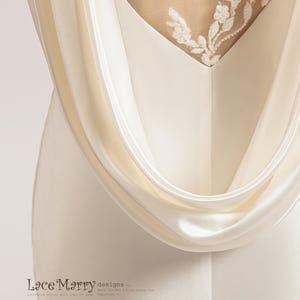 Pu&ograve; includere: Un abito da sposa in satin bianco con schiena scoperta e un delicato ricamo in pizzo. L'abito ha un design fluido e drappeggiato e una silhouette semplice ed elegante. Il testo "Lace Marry designs by Mary Jin Wei & Tina Guang Hua Copyright &copy;" &egrave; stampato sull'abito.