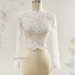 May include: A white lace top with long sleeves and a high neckline. The intricate floral lace pattern covers the entire top, creating a delicate and elegant look. The top is displayed on a mannequin.