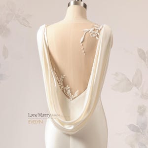 Pu&ograve; includere: Un abito da sposa in satin bianco con schiena scoperta e un pannello trasparente con ricami floreali delicati. L'abito ha una schiena drappeggiata a cappa.