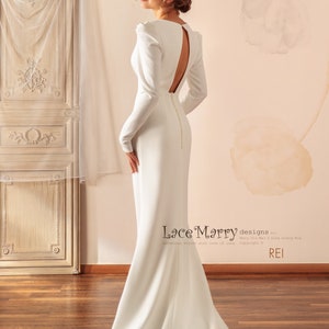 May include: A white, long-sleeved, form-fitting wedding dress with a low back and a train. The dress has a simple, elegant design.