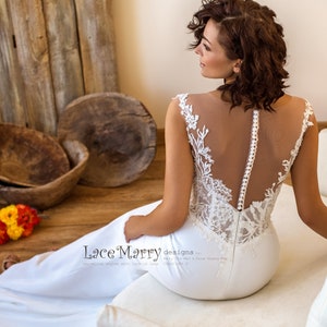 Puede incluir: Un vestido de novia blanco con un corpiño de encaje y una espalda transparente. El vestido tiene una silueta ajustada y una falda fluida. La modelo está sentada en un sofá blanco con un fondo de madera.