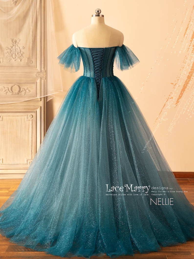 K&ouml;nnte beinhalten: Ein blaues T&uuml;ll-Ballkleid mit einem herzf&ouml;rmigen Ausschnitt und schulterfreien &Auml;rmeln. Das Kleid hat einen Schn&uuml;rr&uuml;cken und ist mit Glitzer verziert. Das Kleid wurde von Lace Marry entworfen und hei&szlig;t "Nellie".