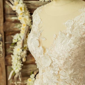 May include: Close-up of a white lace wedding dress with floral embroidery. The dress has a V-neckline and a fitted bodice. The lace is delicate and intricate, with a subtle shimmer.