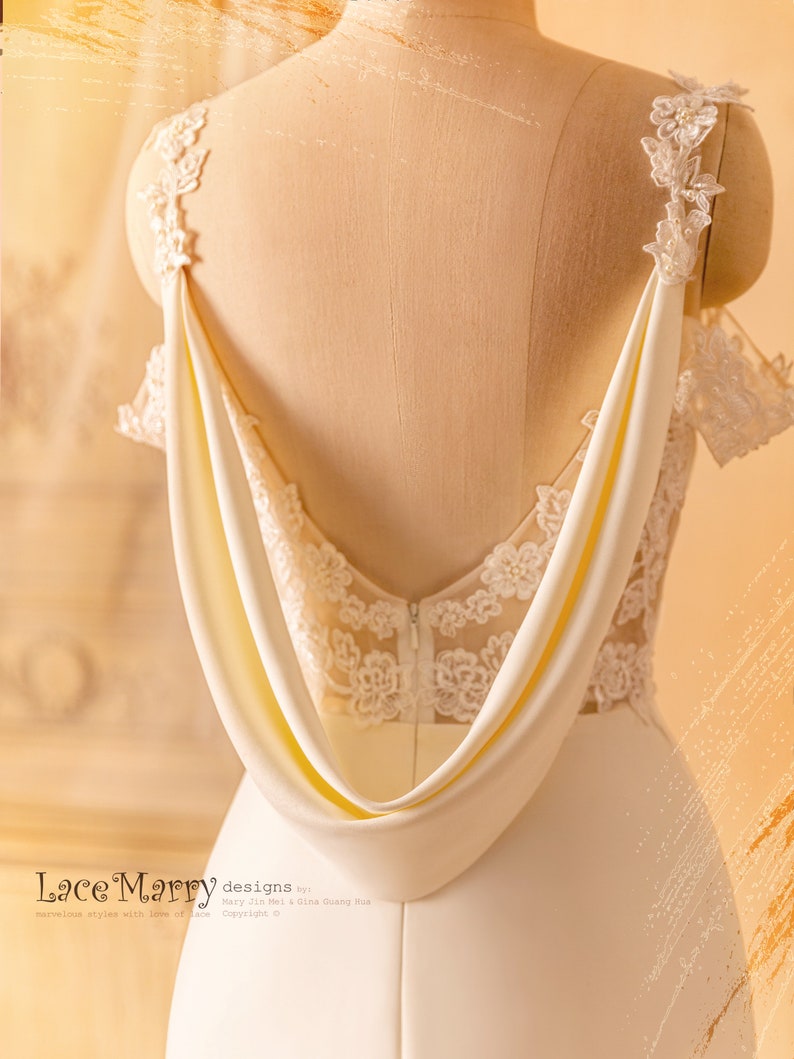 May include: A white wedding dress with a low back and a draped satin detail. The dress features delicate lace appliques on the bodice and straps.