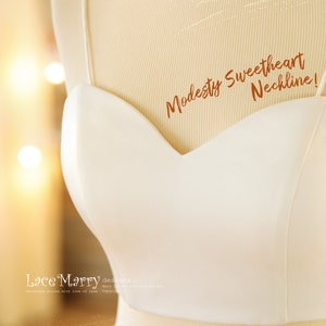 Pu&ograve; includere: Un corpetto bianco con scollatura a cuore su un manichino. Il testo "Modesty Sweetheart Neckline!" &egrave; scritto in marrone sul corpetto. Il testo "Lace Marry designs" &egrave; scritto in nero nella parte inferiore dell'immagine.