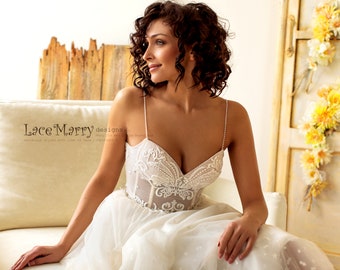 May include: A woman wearing a white wedding dress with a lace bodice and a tulle skirt. The dress has a low back and a sweetheart neckline. The woman is sitting on a white couch and looking to the side. The text "Lace Marry designs" is visible in the image.