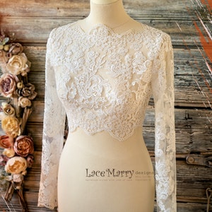 Puede incluir: Bolero de novia blanco de encaje con mangas largas. El encaje tiene un patrón floral y un borde festoneado. El bolero se muestra en un maniquí blanco sobre un fondo de madera.